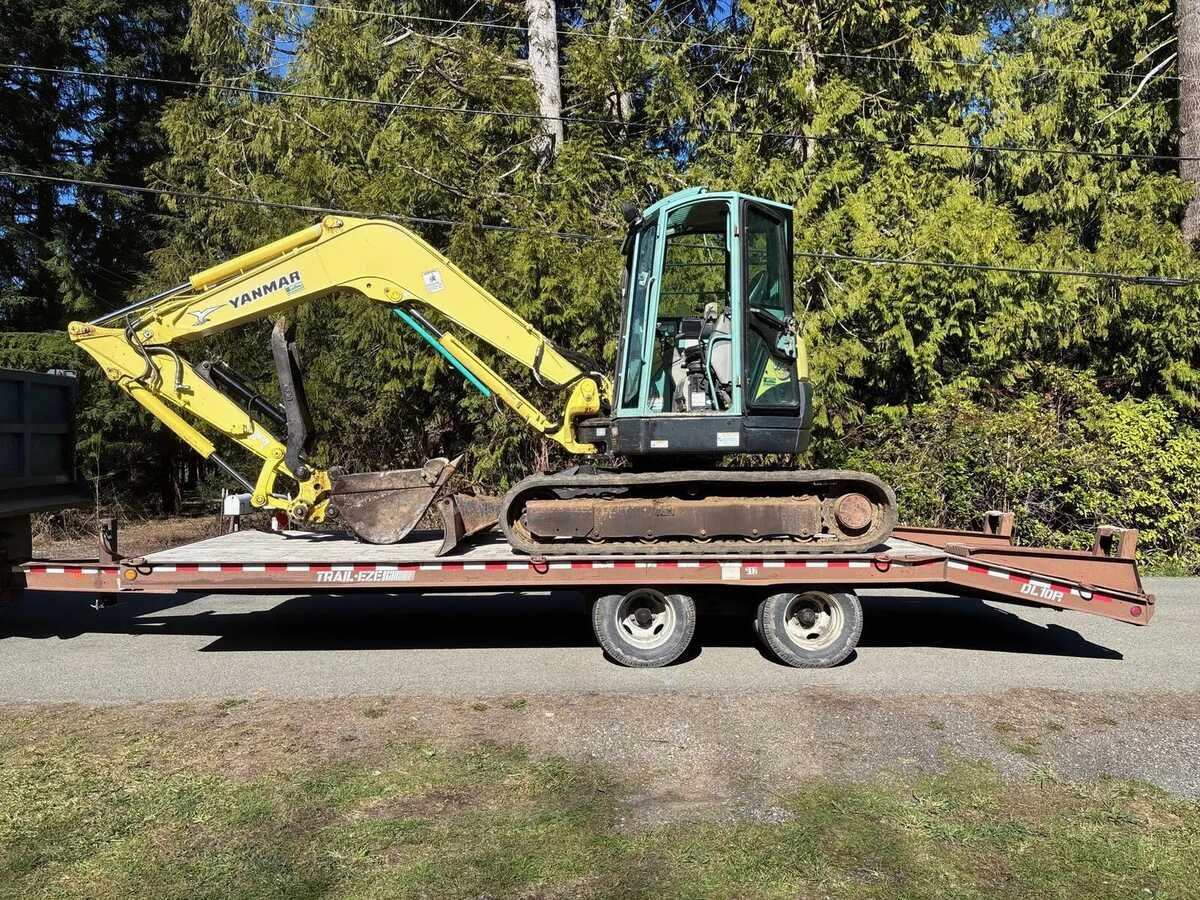 Compact excavator loaded on an equipment trailer beside a timbered lot