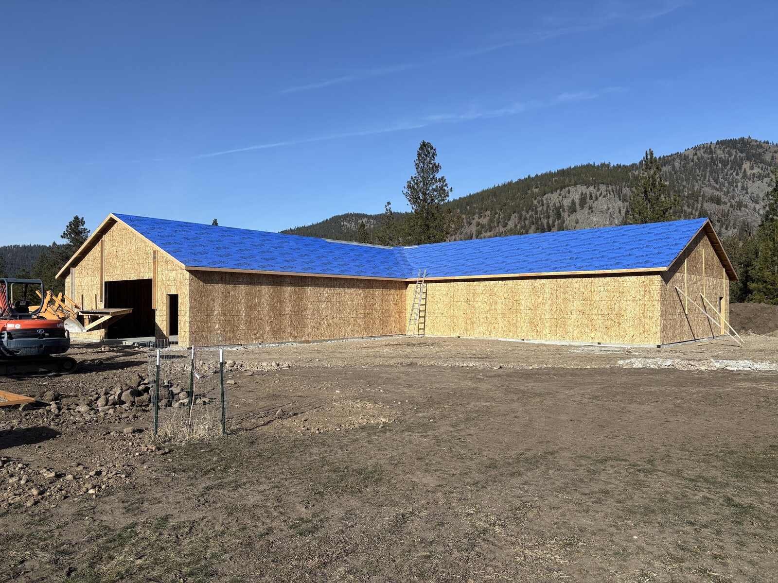 L-shaped pole barn and shop under construction with blue roof underlayment